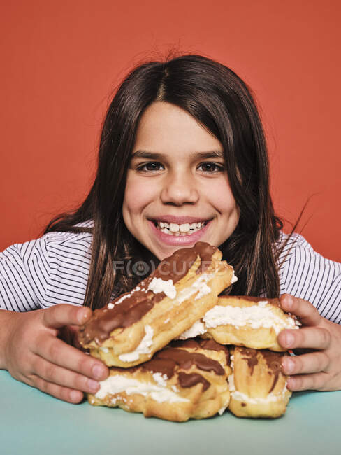 Fröhliches kleines Mädchen in lässiger Kleidung genießt süße Eclairs mit Schokolade und blickt in die Kamera, während sie am Tisch vor rotem Hintergrund sitzt — Stockfoto