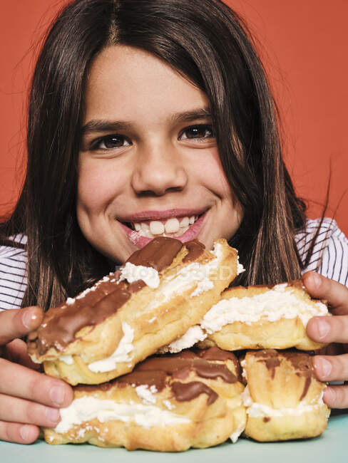 Nahaufnahme Porträt eines fröhlichen kleinen Mädchens in lässiger Kleidung, das süße Eclairs mit Schokolade genießt, während es am Tisch vor rotem Hintergrund sitzt — Stockfoto