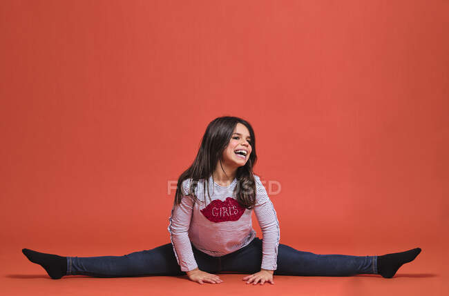 Счастливая девочка-гимнастка в модной одежде смотрит в сторону, сидя с протянутой ногой на красном фоне — стоковое фото