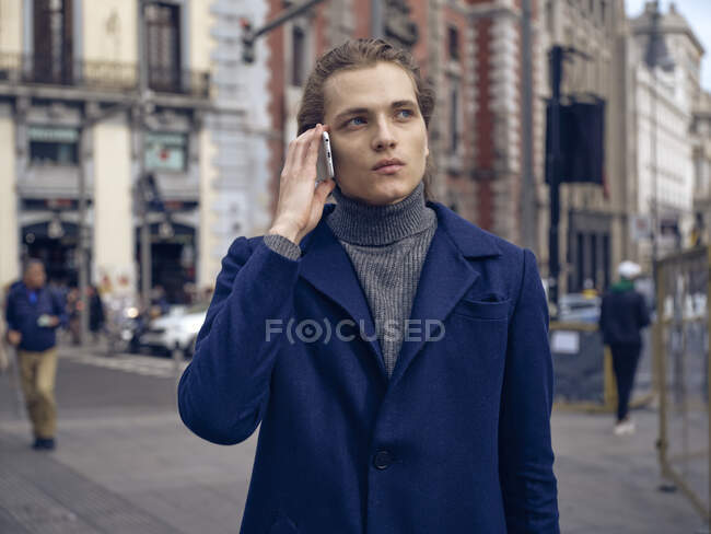 Modern elegant male in trendy coat looking away pensively while having phone conversation on city street — Stock Photo