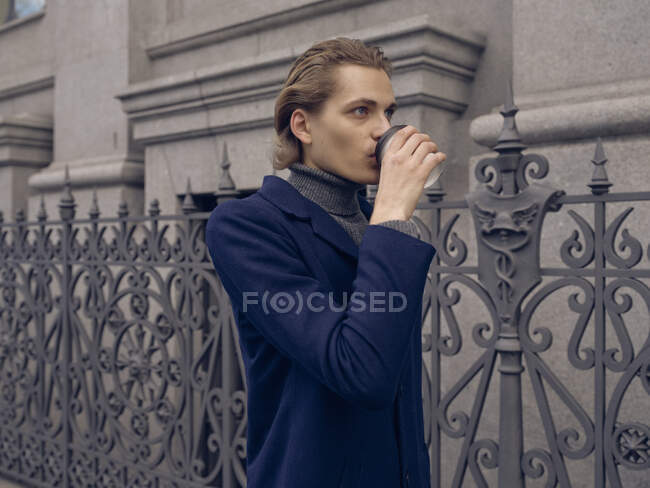 Serious young male with stylish hairdo in trendy coat drinking takeaway coffee while standing near aged metal fence against stone building in city — Stock Photo