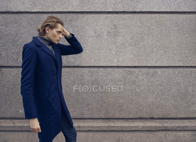 Вид збоку стильного молодого чоловіка з модною зачіскою, одягненою в елегантне пальто, що стоїть на тлі сірої кам'яної стіни в місті — стокове фото