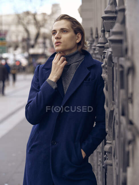 Vista laterale di elegante giovane maschio con acconciatura alla moda vestita in elegante cappotto in piedi contro il muro di pietra grigia in città — Foto stock