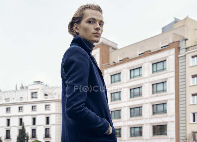Vista lateral do jovem macho elegante com penteado na moda vestido com casaco elegante em pé contra a parede de pedra cinza na cidade — Fotografia de Stock