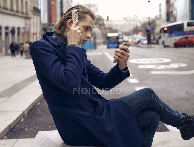 Side view of young handsome male in stylish outfit looking at smartphone and brushing hair while sitting on city street in gray autumn day — Stock Photo