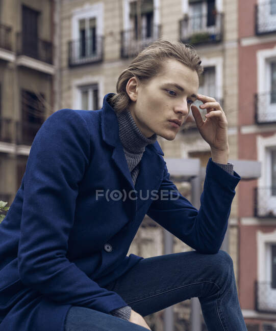 Низкий угол зрения серьезный элегантный молодой мужчина в стильном пальто опираясь на руку и думая, сидя на улице против размытого здания — стоковое фото
