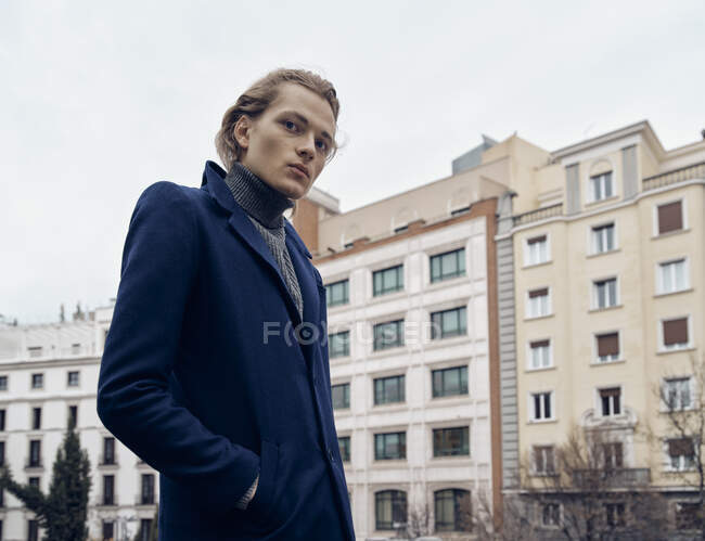 Vista lateral do jovem macho elegante com penteado na moda vestido com casaco elegante em pé contra a parede de pedra cinza na cidade — Fotografia de Stock