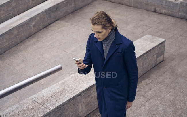 Сверху серьезный молодой бизнесмен в элегантном пальто проверяет сообщение на мобильном телефоне, стоя на каменной лестнице на городской улице — стоковое фото