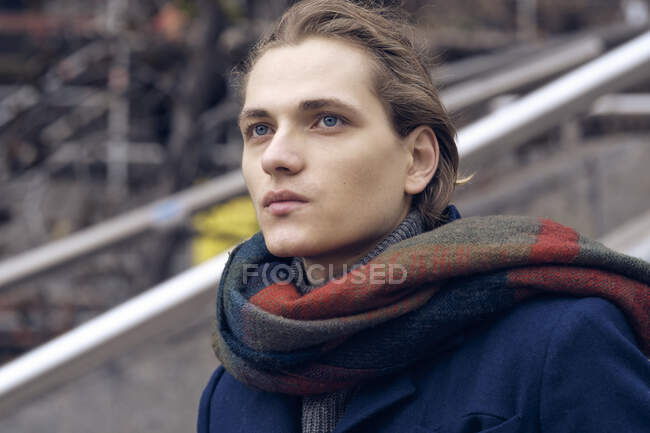 Young handsome confident male in warm coat and scarf looking away while standing against blurred urban background in autumn day — Stock Photo