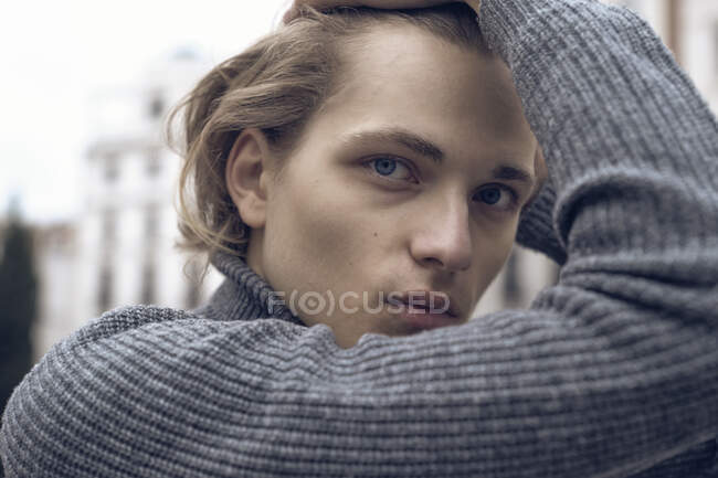 Modern serious young male with stylish haircut in gray warm sweater touching hair and looking at camera while standing on city street — Stock Photo