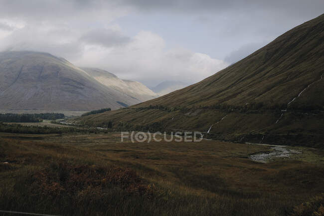 Schottische Landschaft mit schmalem, geschwungenem Fluss, der unter wolkenverhangenem Himmel im Herbst zwischen grasbedeckten Hügeln fließt — Stockfoto