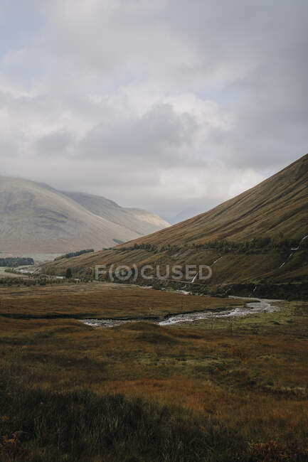 Шотландський краєвид з вузькою вигнутою річкою, вкритою травою пагорбів під хмарним небом восени. — стокове фото