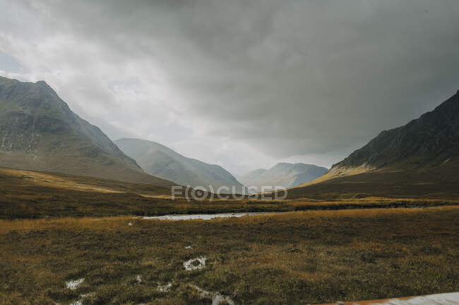 Schottische Landschaft mit schmalem, geschwungenem Fluss, der unter wolkenverhangenem Himmel im Herbst zwischen grasbedeckten Hügeln fließt — Stockfoto