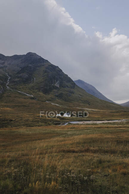 Спокойный пейзаж шотландской сельской местности с желтыми лугами и одинокий домик расположен возле скалистой горы против облачного неба — стоковое фото
