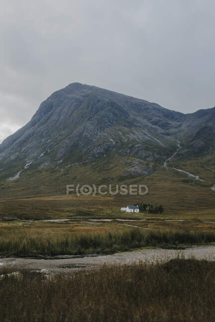 Paisagem tranquila da paisagem rural escocesa com pastagens amarelas e pequena casa solitária localizada perto da montanha rochosa e pequeno rio contra o céu nublado — Fotografia de Stock