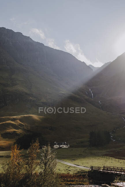 Спокойный пейзаж шотландской сельской местности с желтыми лугами и одинокий домик расположен возле скалистой горы против облачного неба — стоковое фото
