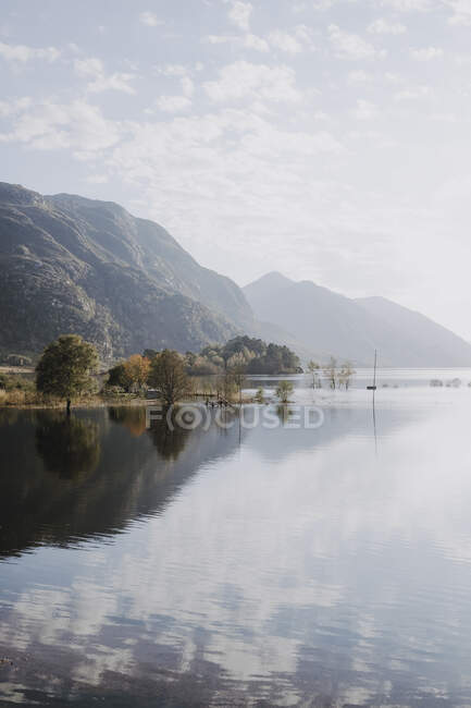 Cenário pitoresco de lago calmo cercado por montanhas rochosas refletidas na água em dia ensolarado com céu nublado na Escócia — Fotografia de Stock