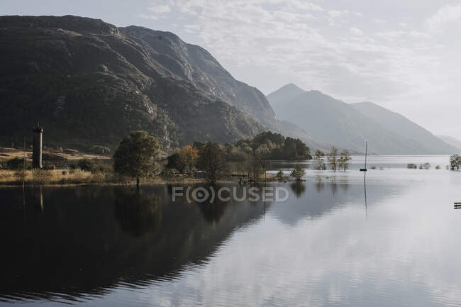 Paysage pittoresque d'un lac calme entouré de montagnes rocheuses reflétées dans l'eau par temps ensoleillé avec ciel nuageux en Écosse — Photo de stock