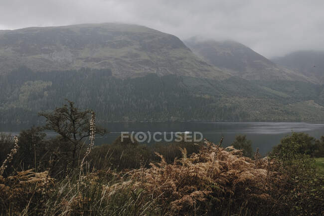 Paisaje brumoso de cordillera cubierta de niebla y nubes cerca de un lago tranquilo en la sierra escocesa - foto de stock