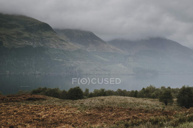 Paysage brumeux d'une chaîne de montagnes couvertes de brouillard et de nuages près d'un lac calme dans les hautes terres écossaises — Photo de stock