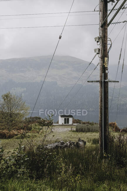 Gloomy Scottish countryside landscape with lonely white house located near mountain lake in overcast day with electric pole and cable in foreground — Stock Photo