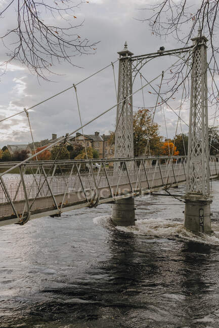 Old suspension bridge crossing river in ancient Scottish city Inverness against gray cloudy sky in autumn day — Stock Photo
