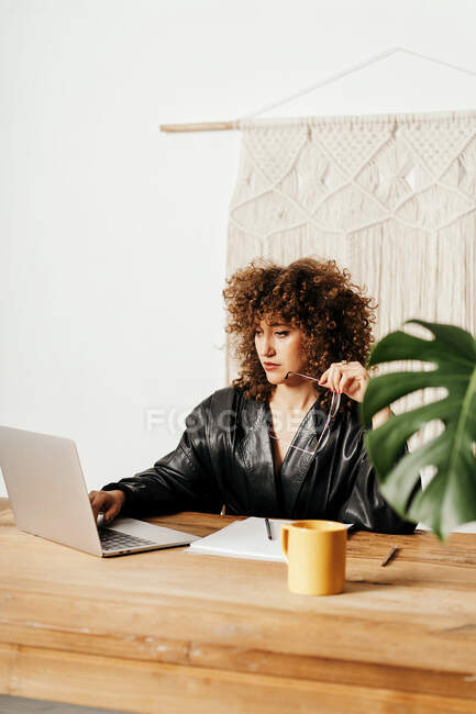 Adult lady in leather jacket and with curly hair sitting at table against macrame decoration and reading data on laptop during work on project in workplace — Stock Photo