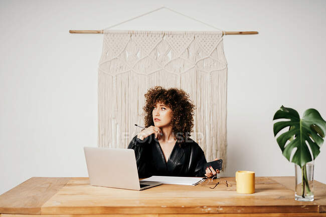 Adult lady in leather jacket and with curly hair sitting at table against macrame decoration and reading data on laptop during work on project in workplace — Stock Photo