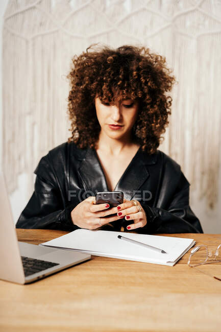Mujer de negocios retro positiva con el pelo rizado sentado en la mesa y el uso de smartphone en la oficina - foto de stock