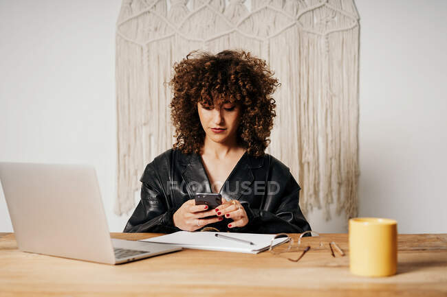 Femme d'affaires rétro positive aux cheveux bouclés assise à table et utilisant un smartphone au bureau — Photo de stock