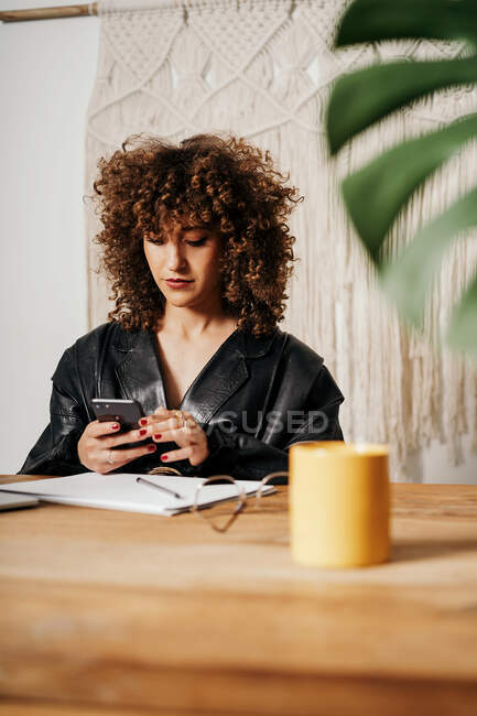 Mujer de negocios retro positiva con el pelo rizado sentado en la mesa y el uso de smartphone en la oficina - foto de stock