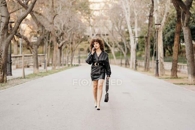 Femme entrepreneur en tenue vintage marchant sur le chemin de l'asphalte et ayant une conversation smartphone tout en se rendant au travail à travers le parc de la ville — Photo de stock