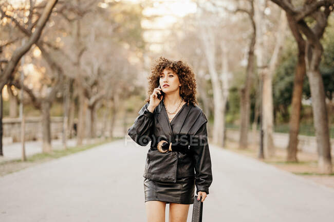 Full body female entrepreneur in vintage outfit walking on asphalt path and having smartphone conversation while commuting to work through city park — Stock Photo