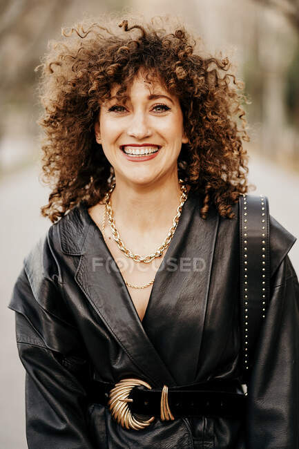 Optimistische Unternehmerin in trendiger Lederjacke und mit lockigem Haar, die auf verschwommenem Straßenhintergrund in die Kamera lächelt — Stockfoto