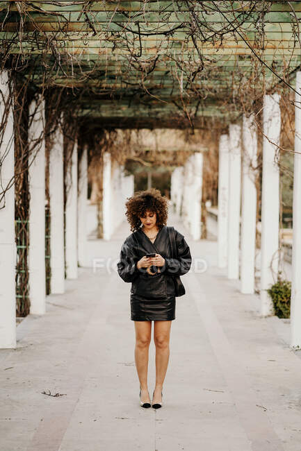 Joven empresaria en chaqueta de cuero negro retro y falda de navegación smartphone mientras está de pie en arco pasaje en el parque antes del trabajo - foto de stock