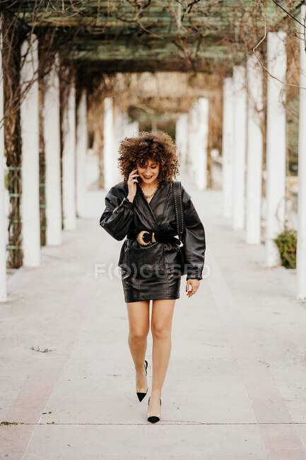 Imprenditrice donna a corpo pieno in abito vintage che cammina su un sentiero asfaltato e fa conversazione con lo smartphone mentre si reca al lavoro nel parco cittadino — Foto stock