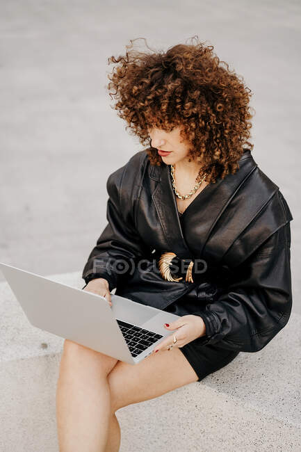 Von oben sieht man eine Geschäftsfrau im schwarzen Lederkostüm, die am Rand sitzt und auf ihrem Laptop Daten durchsucht, während sie an einem Remote-Projekt in der City Street arbeitet. — Stockfoto