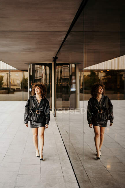 Жіночий менеджер у чорній шкіряній спідній сукні, озираючись, ідучи біля будинку зі скляною стіною на вулицях міста. — стокове фото