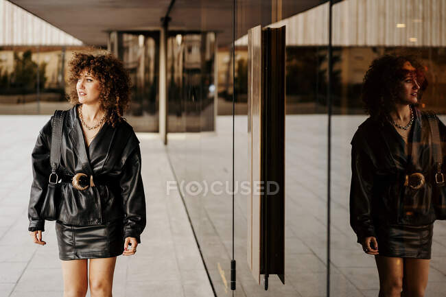 Female manager in black leather skirt suit looking away while walking near building with glass wall on city street — Stock Photo