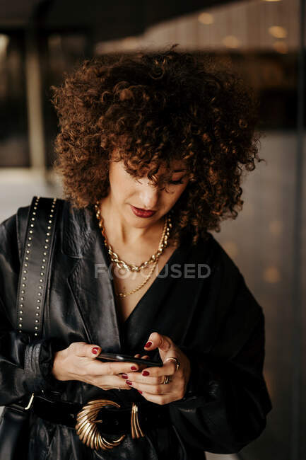 Smart businesswoman in black leather suit and with curly hair looking away and answering phone call while standing near building with glass wall on city street — Stock Photo