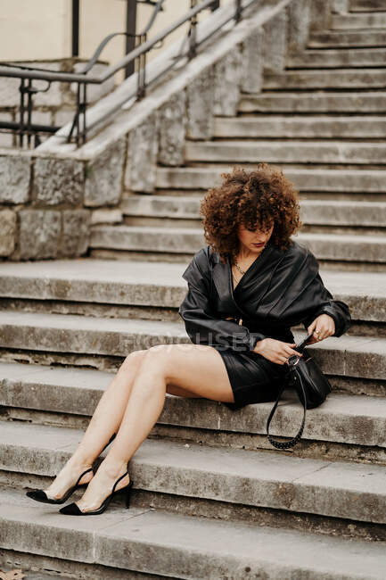 Full length slim female entrepreneur in leather skirt and jacket sitting on concrete steps and browsing smartphone on city street — Stock Photo
