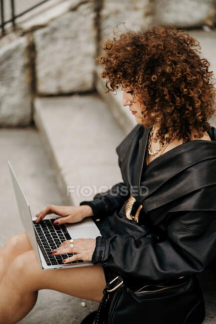 Femme d'affaires habile et bouclée portant costume en cuir noir et veste tapant sur le clavier de l'ordinateur portable tout en étant assis sur les escaliers et travaillant sur un projet à distance sur la rue de la ville — Photo de stock