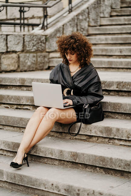 Clevere, lockige Geschäftsfrau in schwarzem Lederanzug und Jacke tippt auf Laptop-Tastatur, während sie auf der Treppe sitzt und an einem Projekt in der City arbeitet — Stockfoto