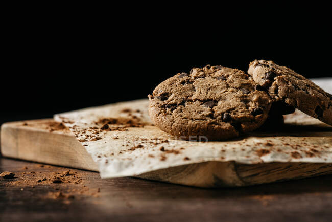 Вкусное домашнее хрустящее печенье с шоколадной крошкой подается на выпечке бумаги на деревянной доске на черном фоне — стоковое фото
