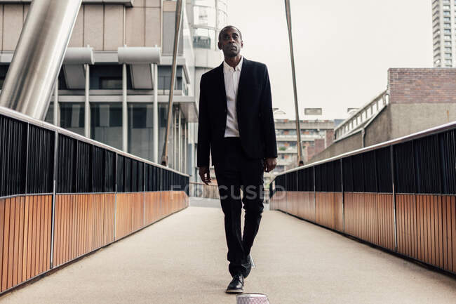 Selbstbewusster afroamerikanischer Mann in elegantem Anzug läuft auf Brücke, während er zur Arbeit auf die Straße der Stadt pendelt — Stockfoto