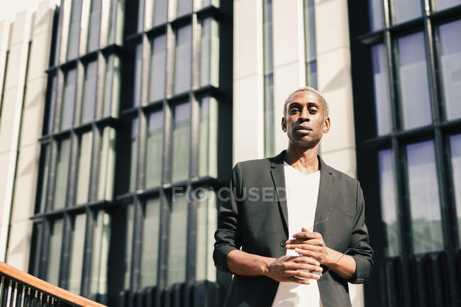 Low angle of confident African American male in trendy suit carrying sunglasses and looking at camera while standing outside modern building on city street — Stock Photo