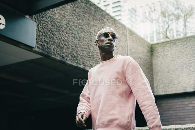Low angle of confident black guy in stylish pink sweatshirt and sunglasses looking away and walking near underground entrance on city street — Stock Photo