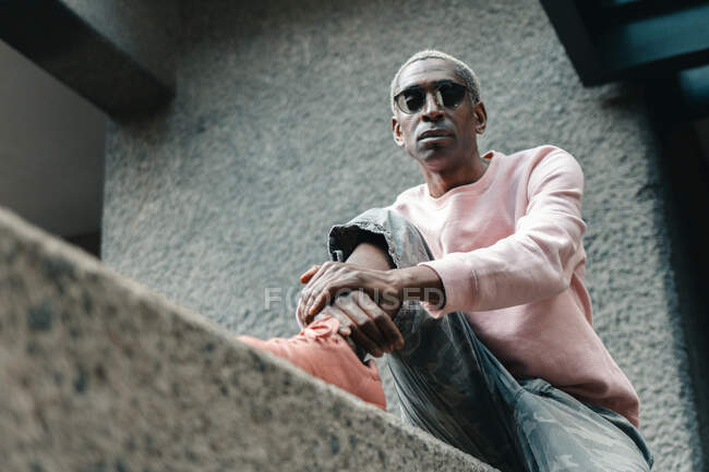 Desde abajo hombre afroamericano adulto en traje de moda y gafas de sol sentado en la frontera de hormigón fuera de edificio moderno en la calle de la ciudad - foto de stock