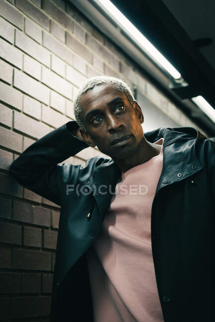 Trendy black male in casual outfit leaning on wall and looking away in illuminated corridor — Stock Photo