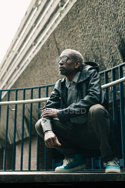 Hombre afroamericano adulto de cuerpo entero con atuendo de moda y gafas de sol sentado en haunches y mirando hacia otro lado fuera del edificio en la calle de la ciudad - foto de stock